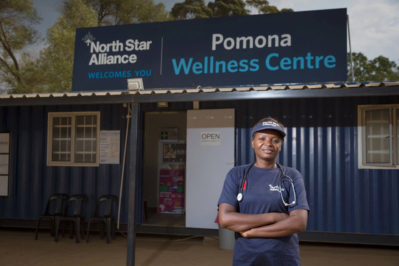 A woman with a stethoscope stands in front of a blue box clinic that says "North Star Alliance welcomes you" and "Pomona Wellness Centre"